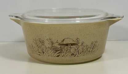 A vintage Pyrex casserole dish with a clear glass lid, featuring a speckled beige body with a brown mushroom and wildflower pattern, set against a plain light background.