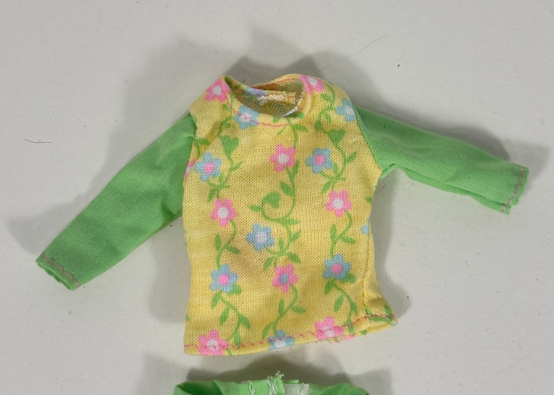 A doll outfit consisting of a long-sleeve top with a yellow floral front panel and green sleeves, paired with matching green bottoms partially visible at the bottom of the frame, laid flat on a light gray surface.
