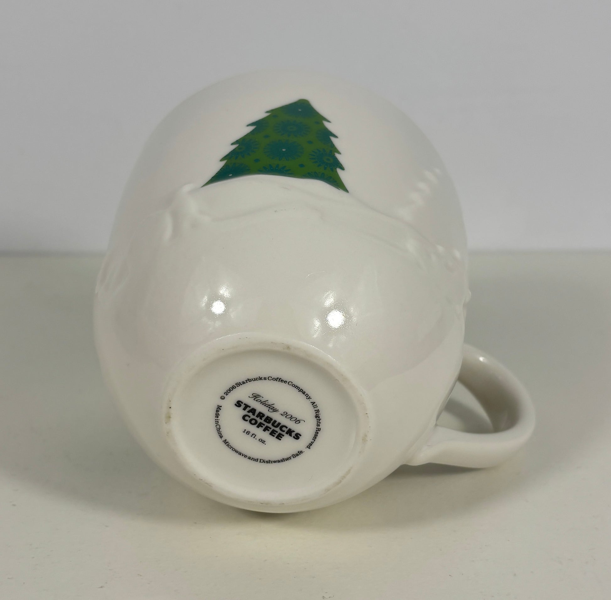 A white ceramic Starbucks coffee mug shown upside down, displaying a green Christmas tree design on the side and the brand's circular logo printed on the base.