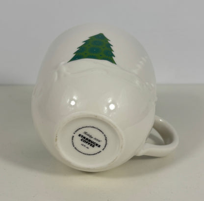 A white ceramic Starbucks coffee mug shown upside down, displaying a green Christmas tree design on the side and the brand's circular logo printed on the base.