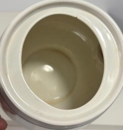 Close-up of the inside of a glossy ceramic jar or pot with a matching lid, showing a cream-colored glazed interior and a small portion of a hand holding it at the lower left.
