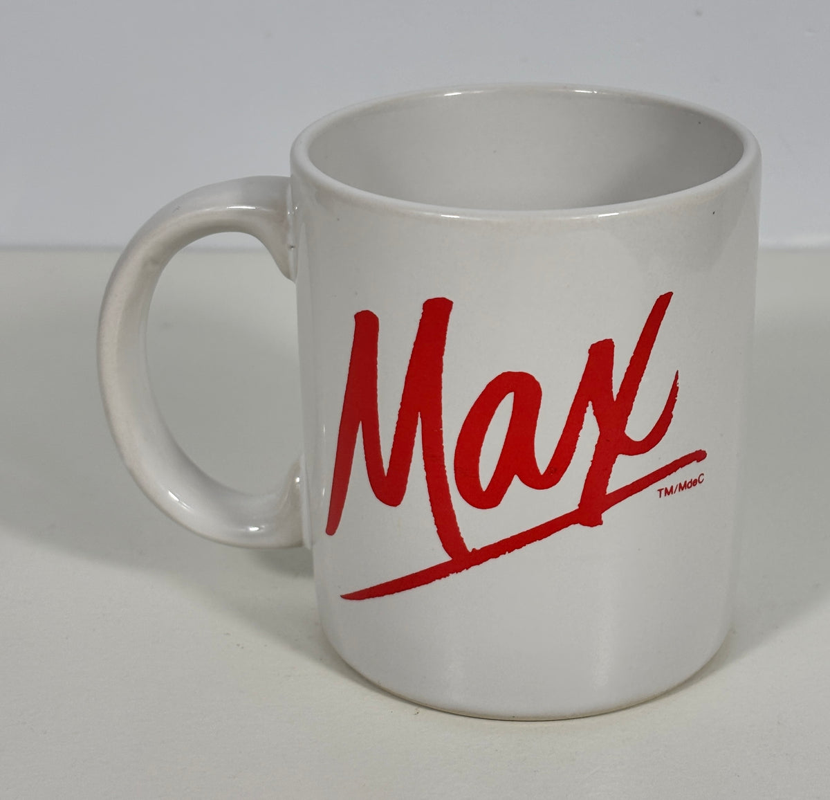 A white ceramic coffee mug featuring the word 'Max' printed in a red cursive script, with a small 'TM-MAC' trademark mark below it, set against a plain light background.