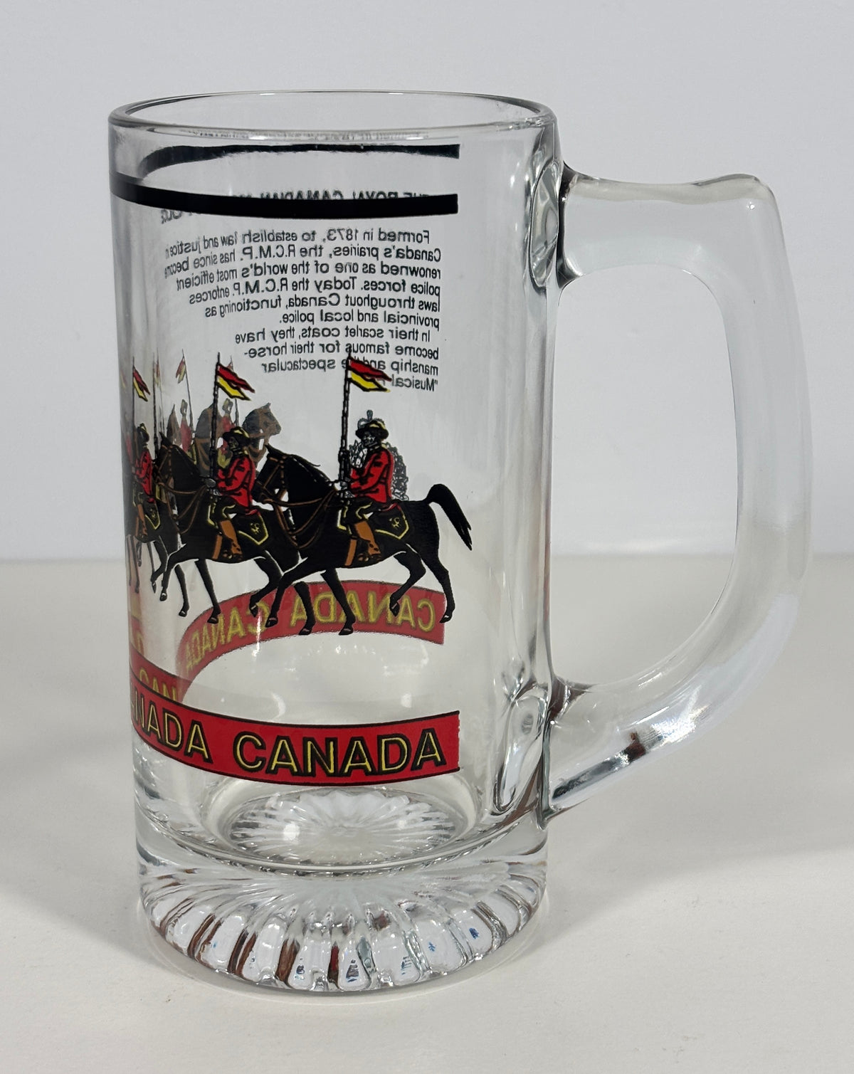 A clear glass beer mug with a handle, featuring a printed graphic of Canadian mounted police riders and the text 'CANADA CANADA' on the front, set against a plain light background.