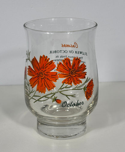 A clear glass candle holder with a vintage-style botanical design featuring orange cosmos flowers and green stems, printed with 'October' and 'Flower of October' text.