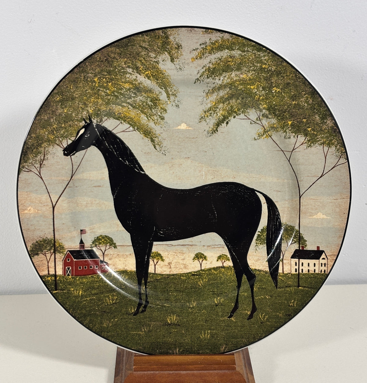 A round decorative plate featuring a black horse in a pastoral landscape with trees, a red barn, and a white house, displayed on a wooden stand against a light gray background.
