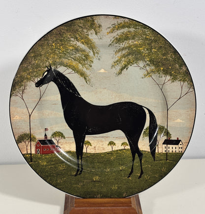 A round decorative plate featuring a black horse in a pastoral landscape with trees, a red barn, and a white house, displayed on a wooden stand against a light gray background.