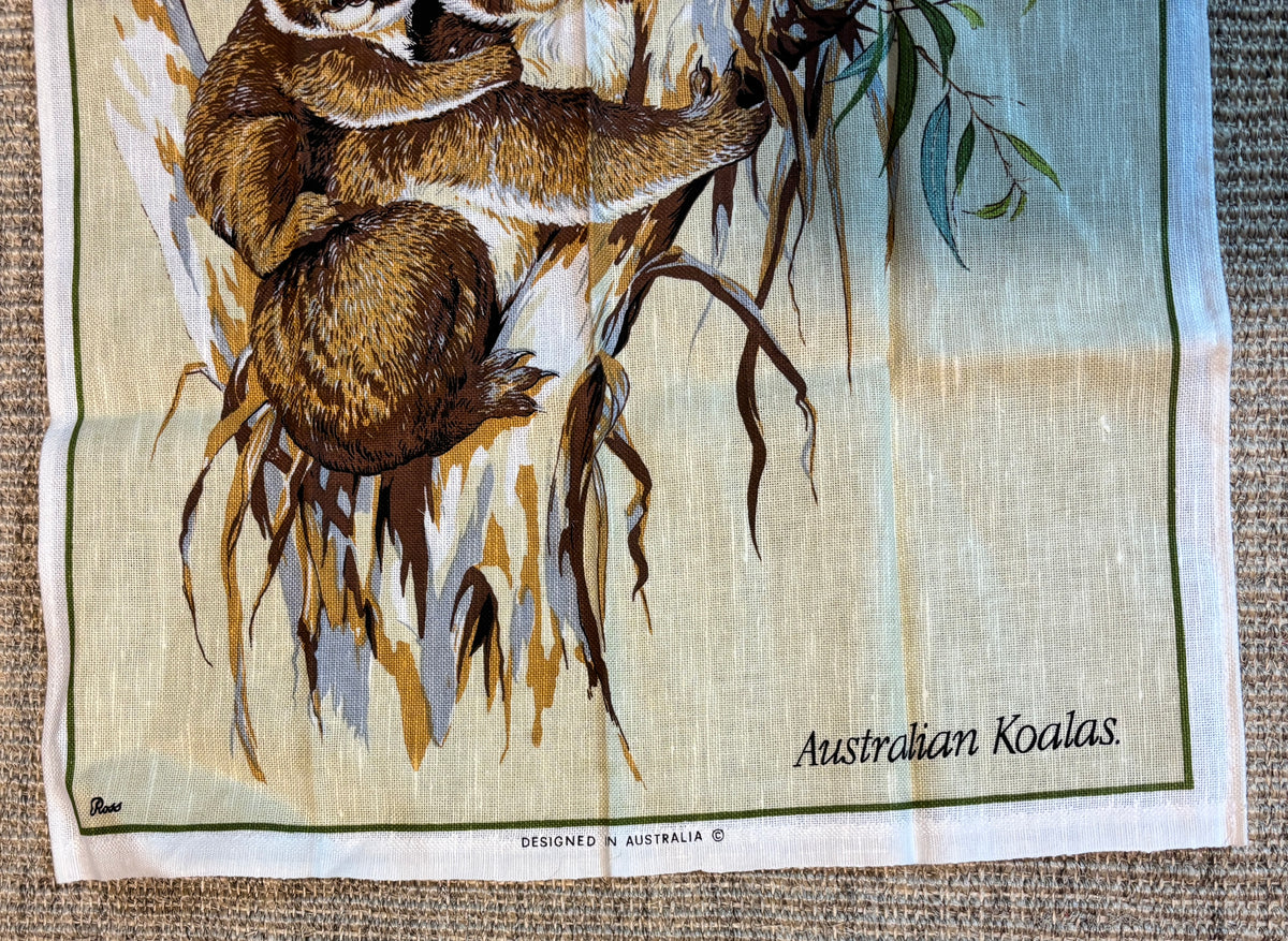 A vintage-style Australian koala art print or textile panel laid flat on a woven surface. The design shows two koalas clinging to a tree branch with leaves, on a pale cream background with green border lines and the text 'Australian Koalas' in the lower right.