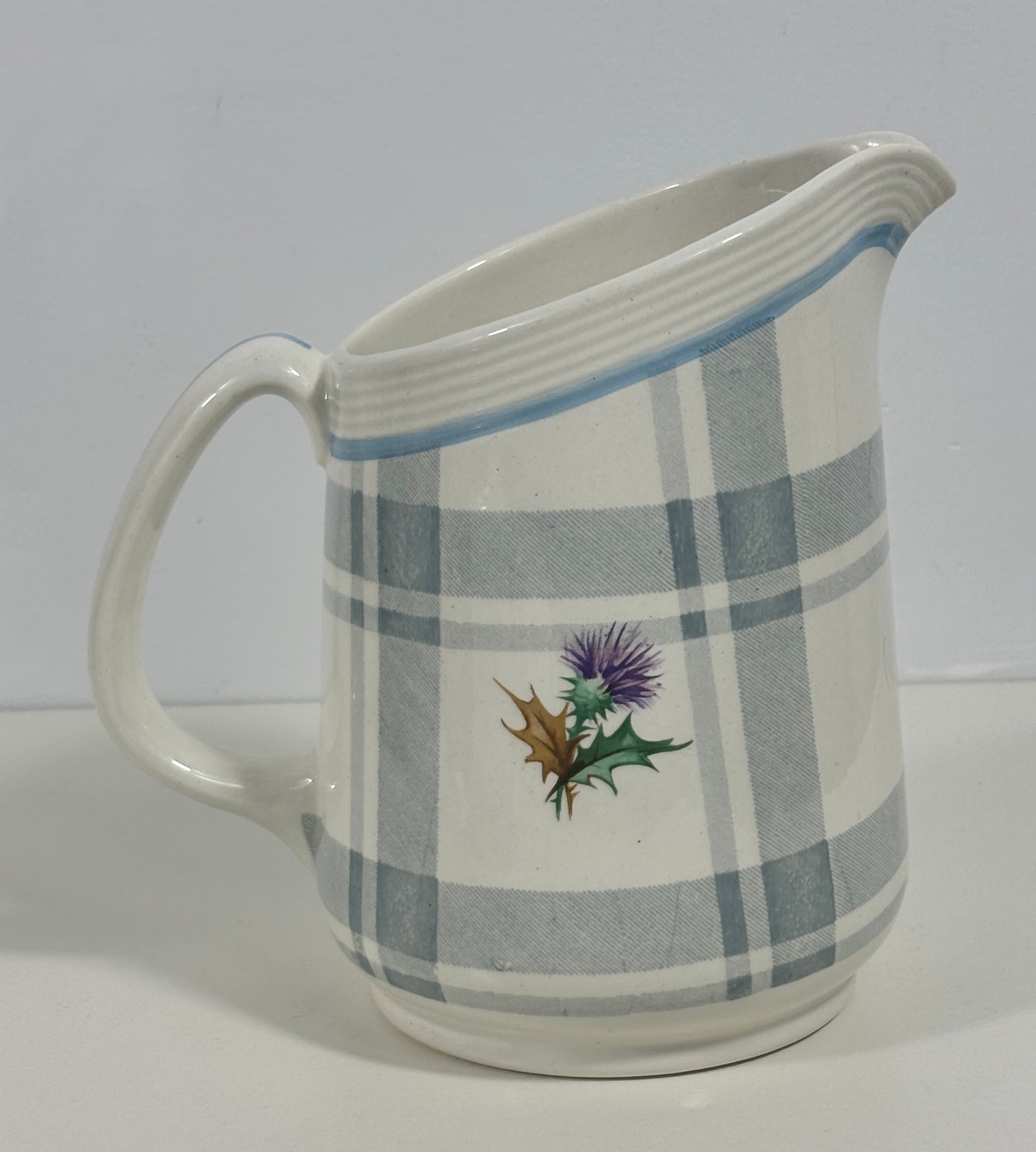 A white ceramic pitcher with a gray and white plaid pattern, featuring a small painted thistle design on the front and a light blue stripe near the rim.
