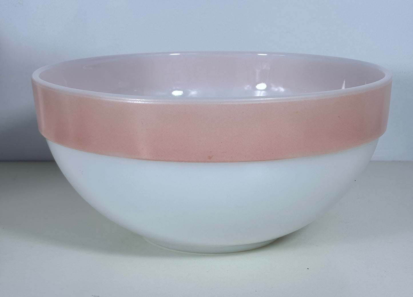 A vintage glass mixing bowl with a white exterior and a dusty pink glazed rim, set against a plain light gray background.