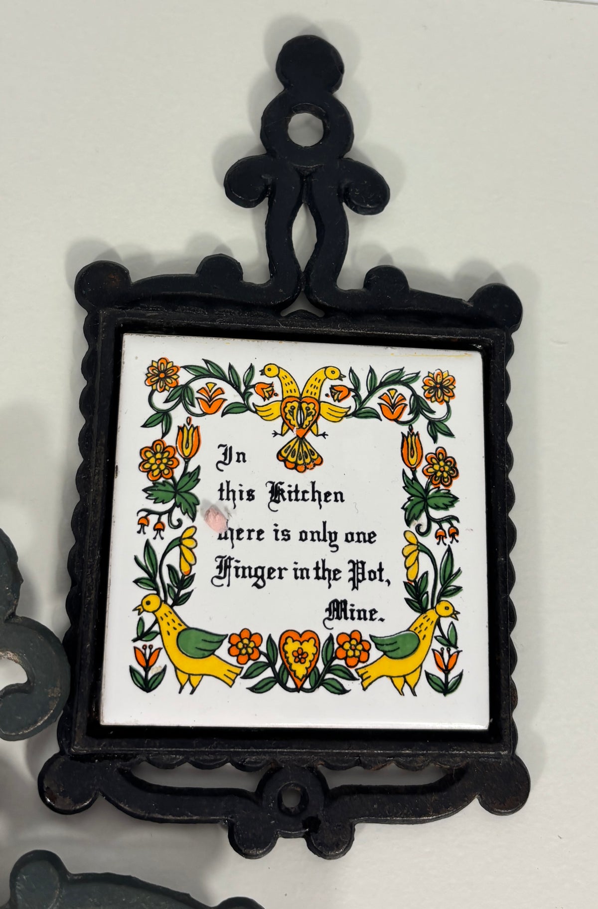 A decorative black metal wall plaque with a white center panel featuring a folk-art floral border and the text, 'In this Kitchen there is only one Finger in the Pot, Mine.'.