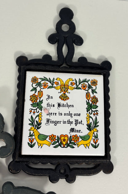 A decorative black metal wall plaque with a white center panel featuring a folk-art floral border and the text, 'In this Kitchen there is only one Finger in the Pot, Mine.'.