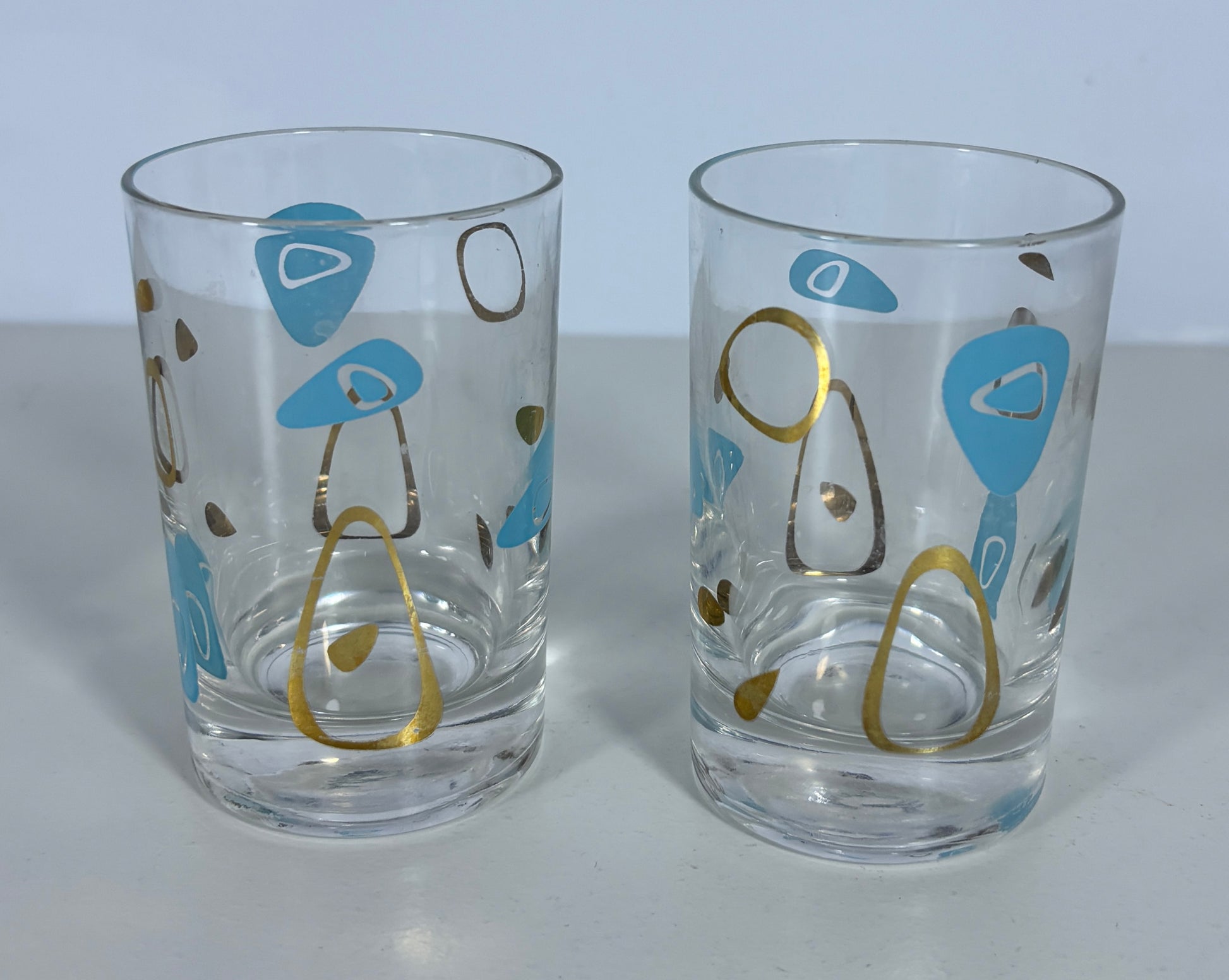 Two clear glass tumblers with mid-century modern abstract patterns in light blue, gold, and brown on a light gray background.