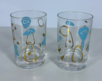 Two clear glass tumblers with mid-century modern abstract patterns in light blue, gold, and brown on a light gray background.