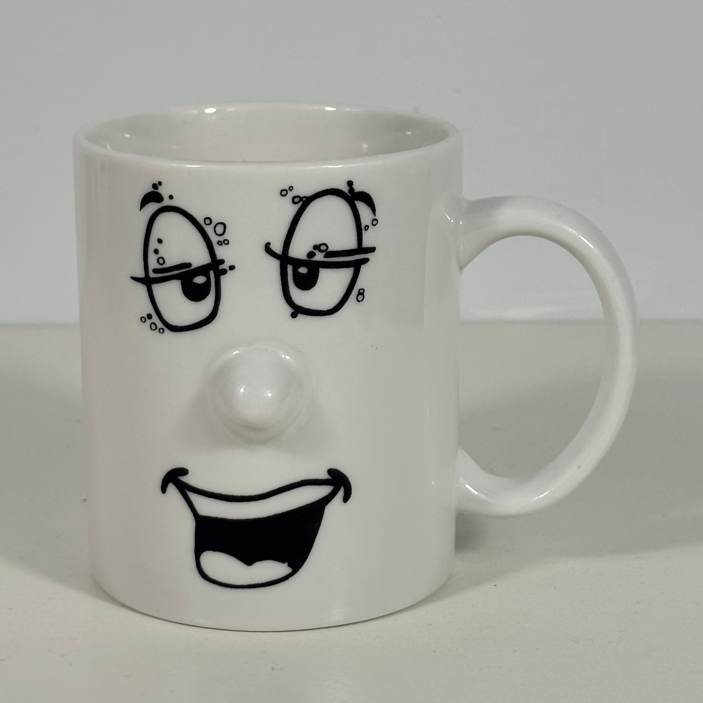 A white ceramic mug featuring a black hand-drawn face design with winking eyes, a raised nose, and a wide open smile.