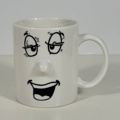 A white ceramic mug featuring a black hand-drawn face design with winking eyes, a raised nose, and a wide open smile.
