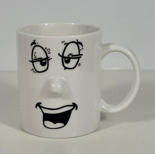 A white ceramic mug featuring a black hand-drawn face design with winking eyes, a raised nose, and a wide open smile.