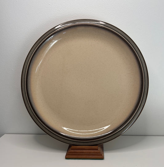 Round ceramic plate with a brownish-beige center and metallic rim on a wooden stand against a white background