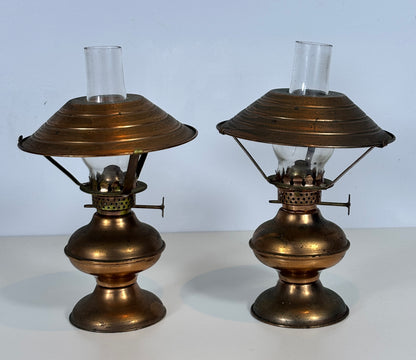 A pair of vintage brass oil lamps with clear glass shades, displayed side-by-side against a light blue background.