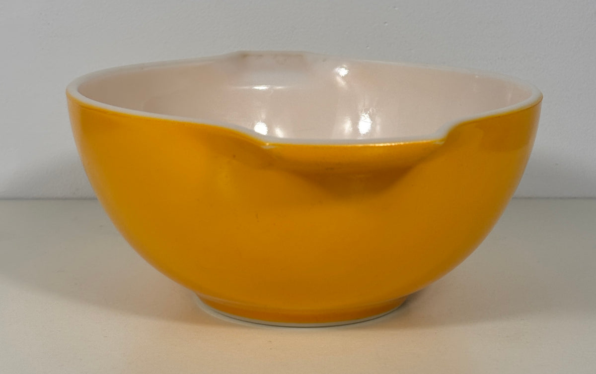 A vintage ceramic mixing bowl with a glossy mustard yellow exterior and a light pinkish-beige interior, featuring a small pouring spout on the side.