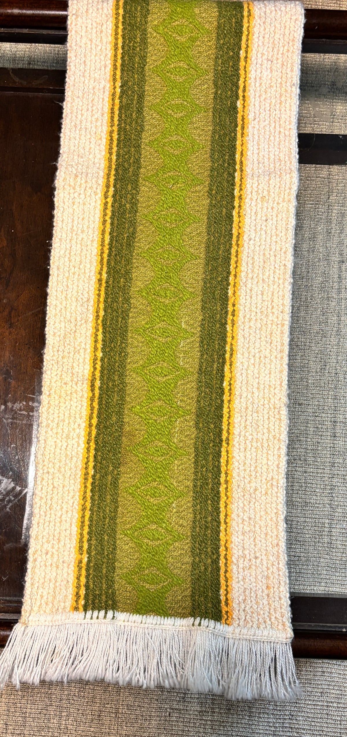 A woven table runner with fringe, featuring vertical stripes in olive green, lime green, and yellow on a cream background, displayed on a dark wooden table.