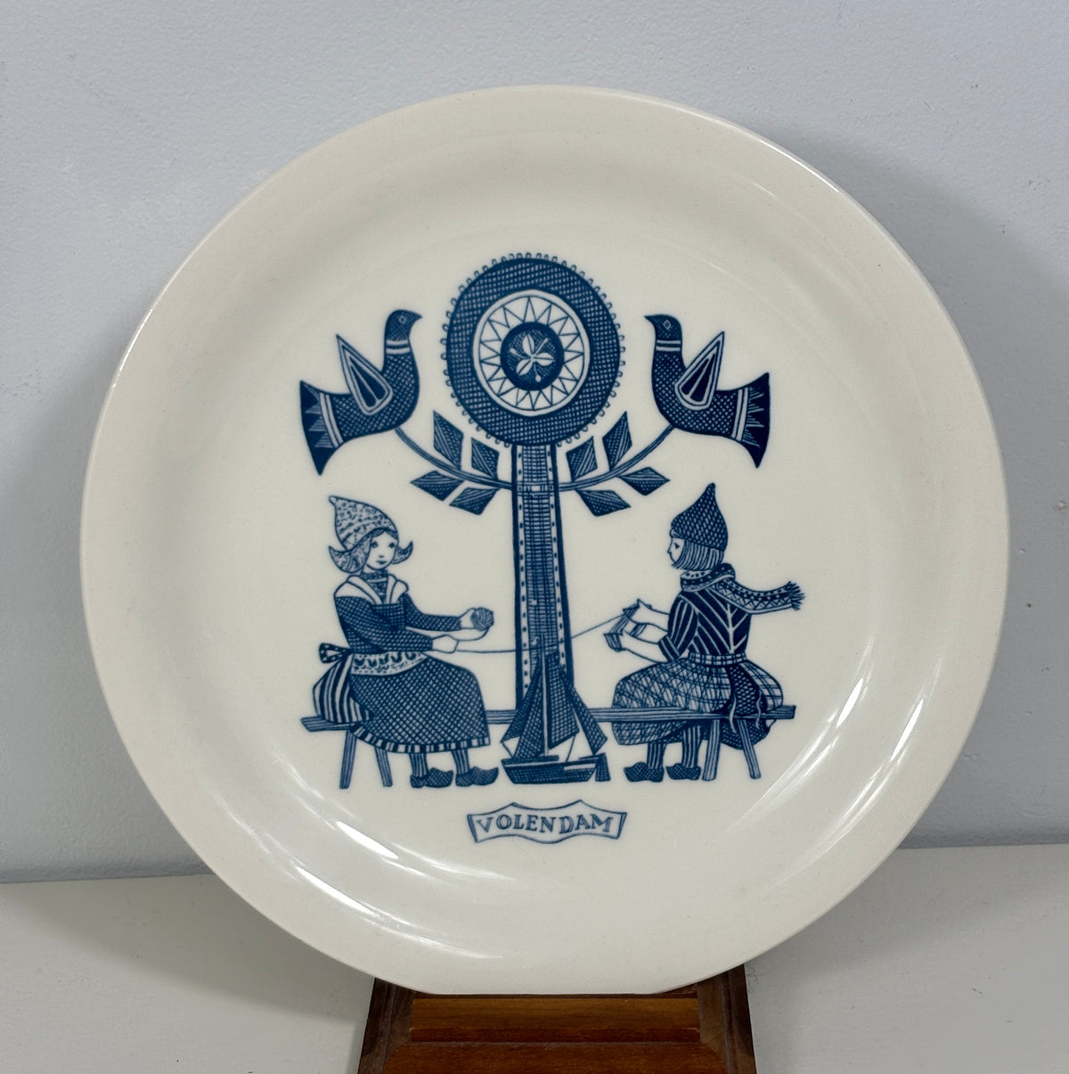 A white ceramic plate with a blue Folkfaktura design, featuring two figures seated under a tree with birds, displayed on a wooden stand against a light gray wall.