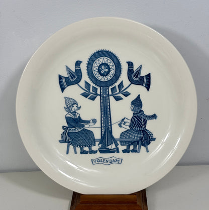 A white ceramic plate with a blue Folkfaktura design, featuring two figures seated under a tree with birds, displayed on a wooden stand against a light gray wall.