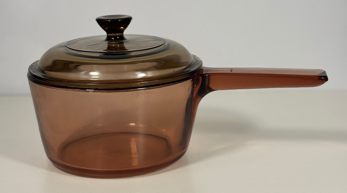 A translucent amber-brown glass saucepan with a matching lid and handle, photographed on a plain light background.