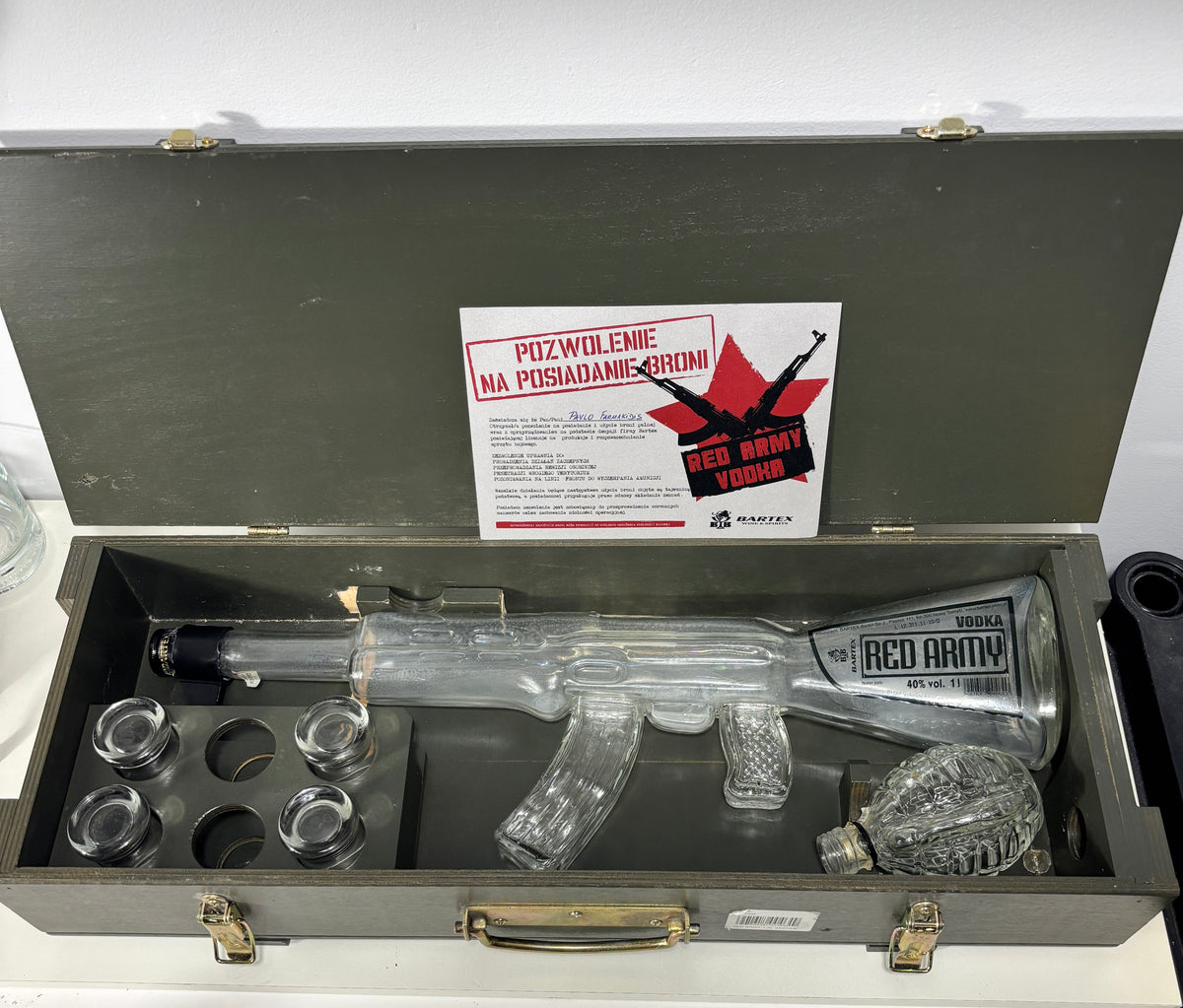 A clear glass replica of an AK-47 rifle with a red and black 'Red Army Vodka' label, presented inside a gray metal case with six small glass jars and a white instruction card.