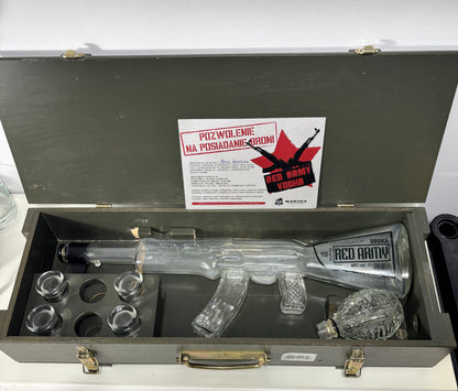 A clear glass replica of an AK-47 rifle with a red and black 'Red Army Vodka' label, presented inside a gray metal case with six small glass jars and a white instruction card.