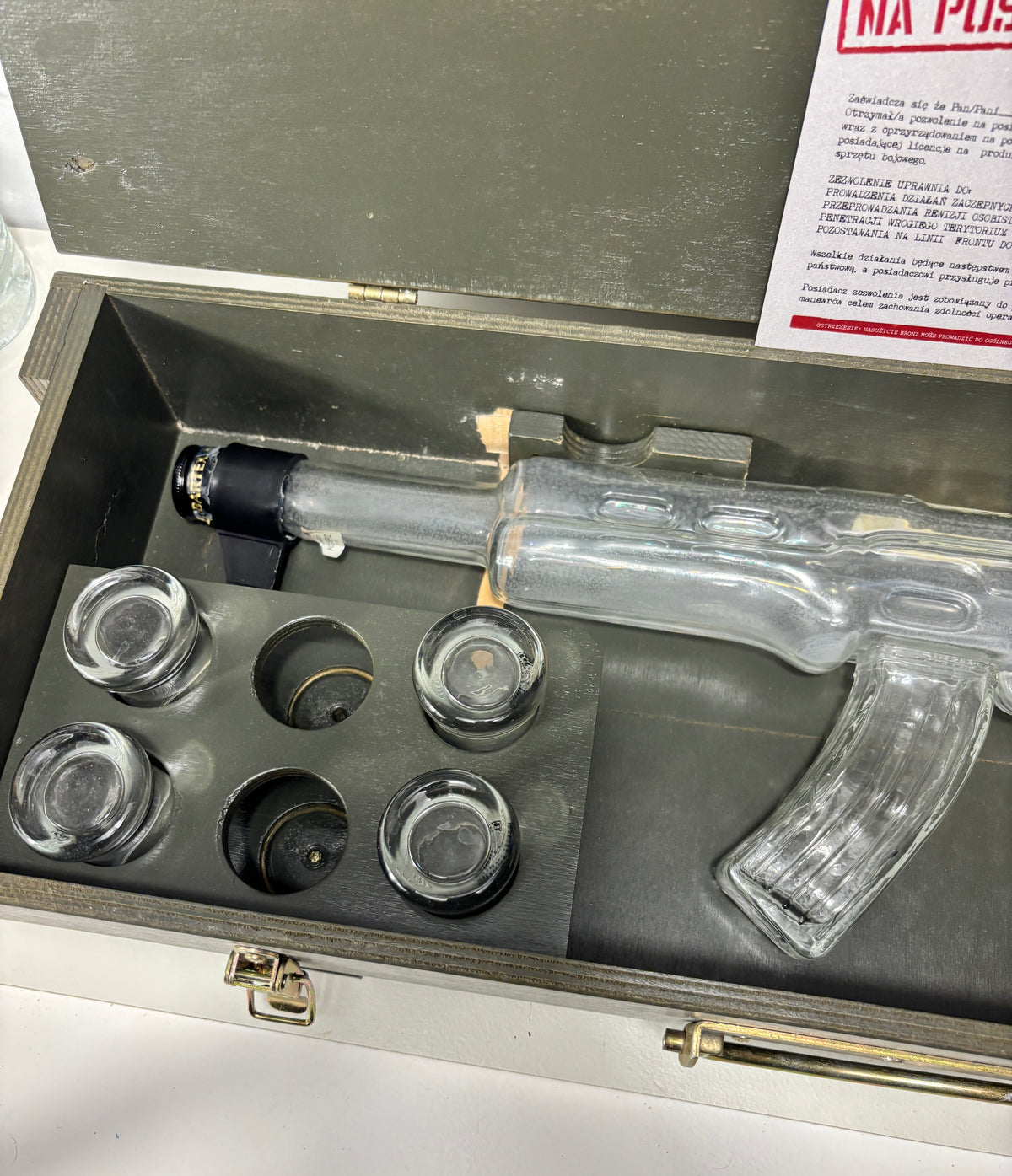 A clear plastic replica of an AK-47 rifle displayed inside an open gray metal case with six clear glass or acrylic bullet holes, alongside a printed instruction card.