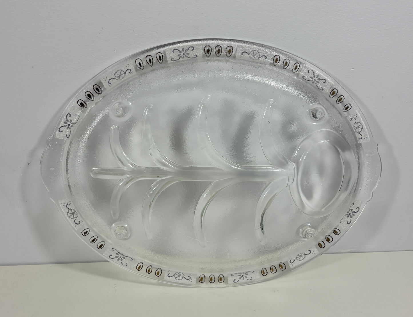 A clear glass serving platter with a leaf-shaped center and decorative border detailing, photographed against a plain light background.