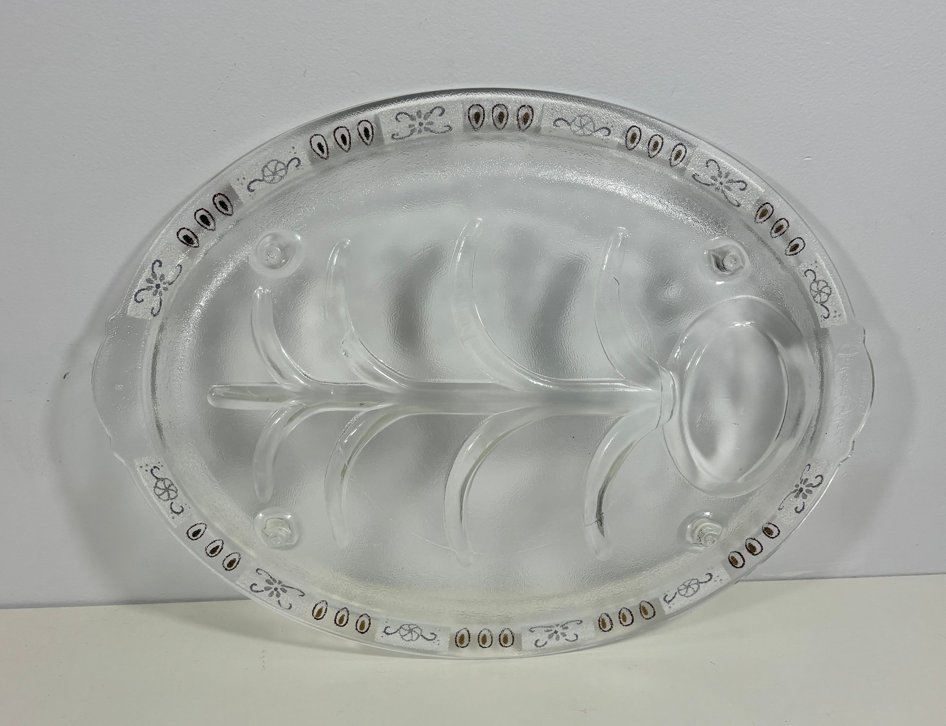 A clear glass serving platter with a leaf-shaped center and decorative border detailing, photographed against a plain light background.
