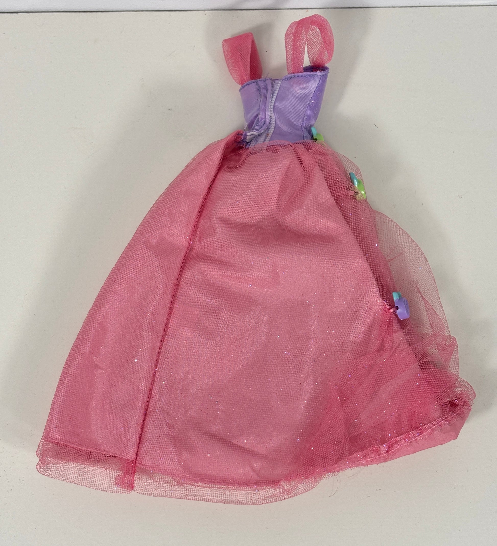 A pink doll dress with a purple bodice and pink tulle straps, laid flat on a light-colored surface.