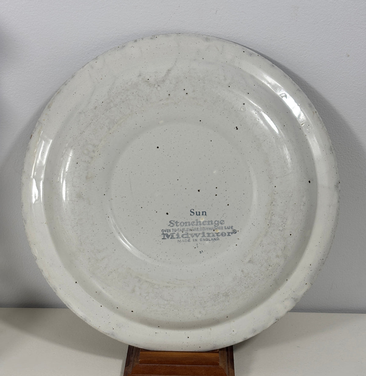 A round white ceramic Stonchase plate with a speckled glaze, displayed on a wooden stand against a light gray wall. The plate features a printed blue label in the center reading 'Sun Stonchase'.