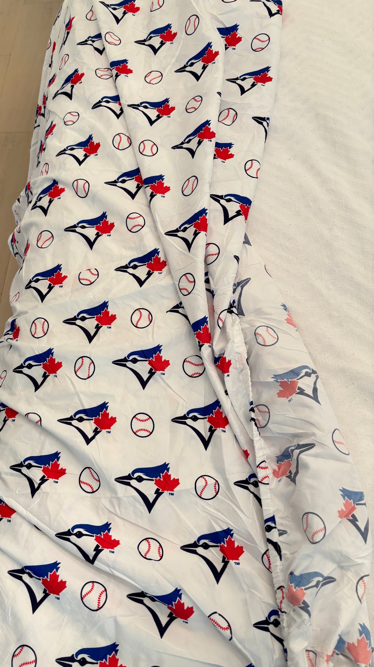 A white bedding cover, printed with a repeating pattern of Toronto Blue Jays baseball logos, baseballs, and maple leaves. The fabric is draped over a bed, showing the texture and print details.