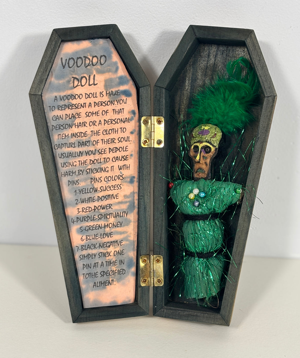A decorative wooden coffin-shaped display with an open hinged lid, showing printed instructions on the left panel and a green cloth-wrapped doll with a feathered headpiece on the right. The product is photographed against a plain light wall.