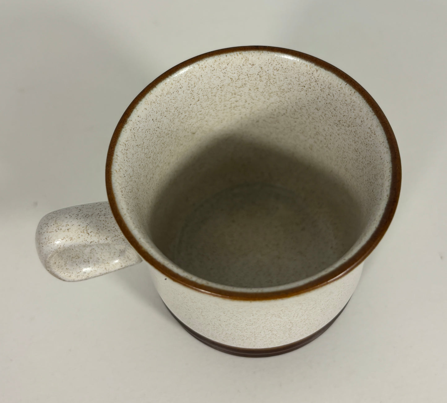 A speckled ceramic mug with a warm off-white interior and exterior, accented by a dark brown rim and base, shown from a top-down angle on a light gray background.