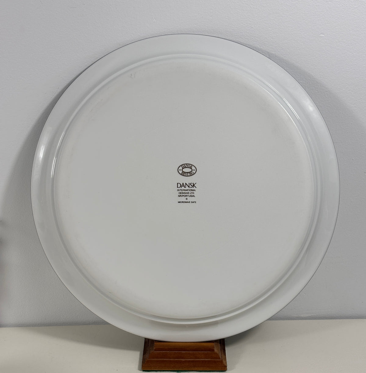 A white round ceramic serving platter or decorative plate with a small black 'DANSK' logo in the center, displayed on a small wooden stand against a light gray wall.