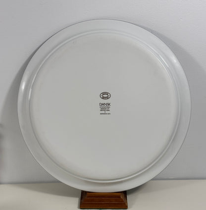 A white round ceramic serving platter or decorative plate with a small black 'DANSK' logo in the center, displayed on a small wooden stand against a light gray wall.