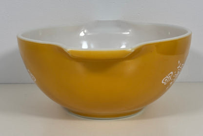 A vintage Pyrex mixing bowl in yellow with a white interior and white printed floral designs on the outside, set against a plain light gray background.