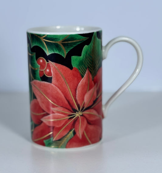 A white ceramic mug decorated with a festive holiday design featuring a large red poinsettia flower, green holly leaves, and red berries on a black background.