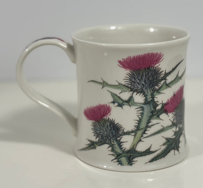 A white ceramic mug featuring a detailed botanical illustration of pink thistles and green spiky leaves, set against a neutral light gray background.