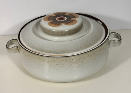A ceramic lidded casserole dish with side handles, featuring a speckled off-white glaze, a dark rim, and a floral design on the lid knob.