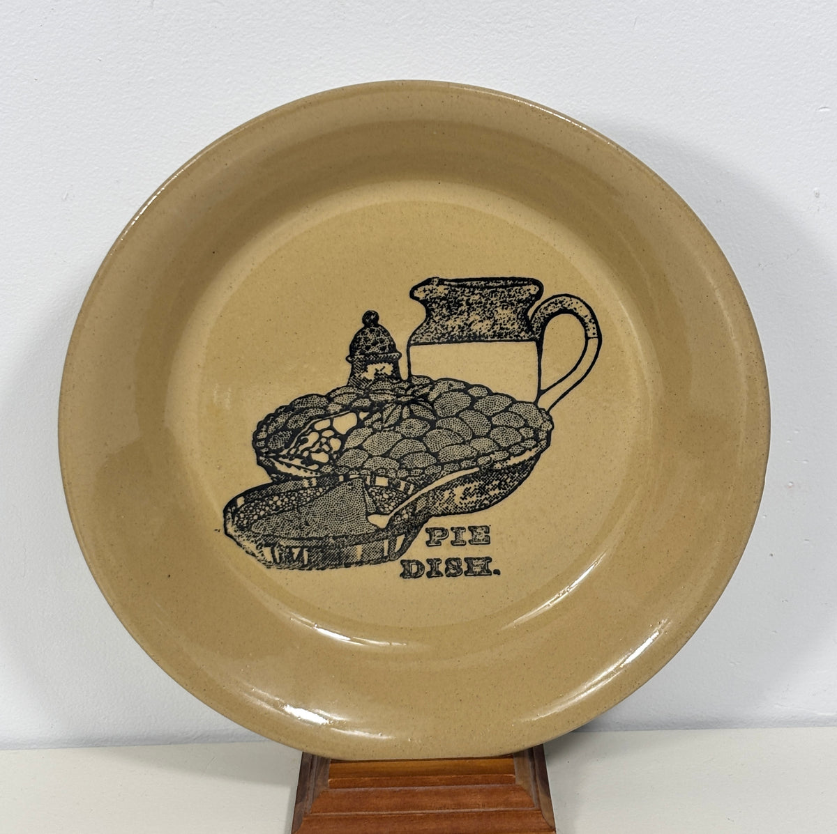 A round beige ceramic pie dish with a black printed illustration of pie, crust, and a pitcher, with the words 'PIE DISH.' on the front, displayed on a wooden stand against a white wall.