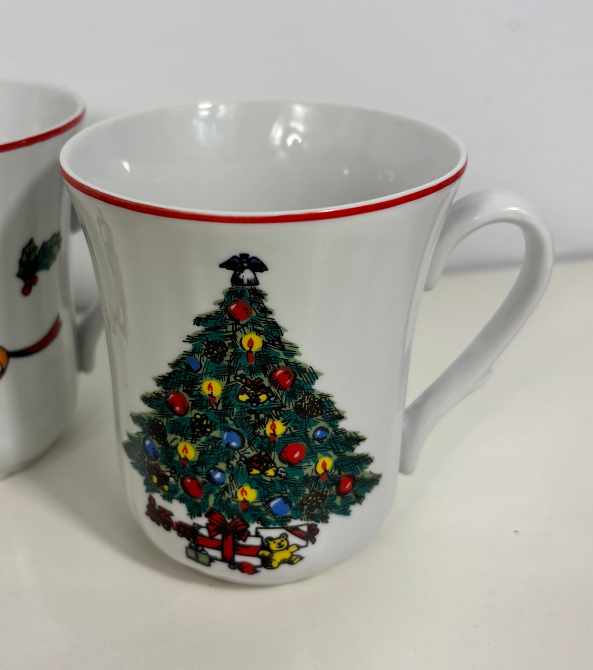 A white ceramic Christmas mug with a red rim, featuring a printed Christmas tree decorated with colorful ornaments and a red gift box underneath. A second similar mug is partially visible on the left edge.