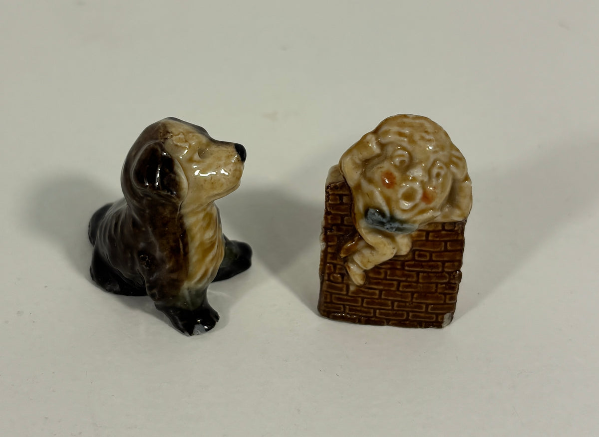 Two small ceramic figurines on a plain light background: a glossy black-and-tan dog on the left and a beige dog sitting on a brown basket with a blue bow tie on the right.