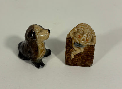 Two small ceramic figurines on a plain light background: a glossy black-and-tan dog on the left and a beige dog sitting on a brown basket with a blue bow tie on the right.