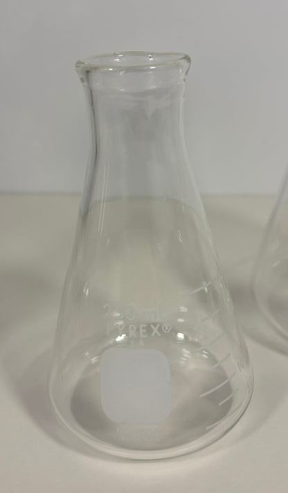 A clear glass Erlenmeyer flask with etched measurement markings and a frosted square label on the front, photographed on a light neutral background.
