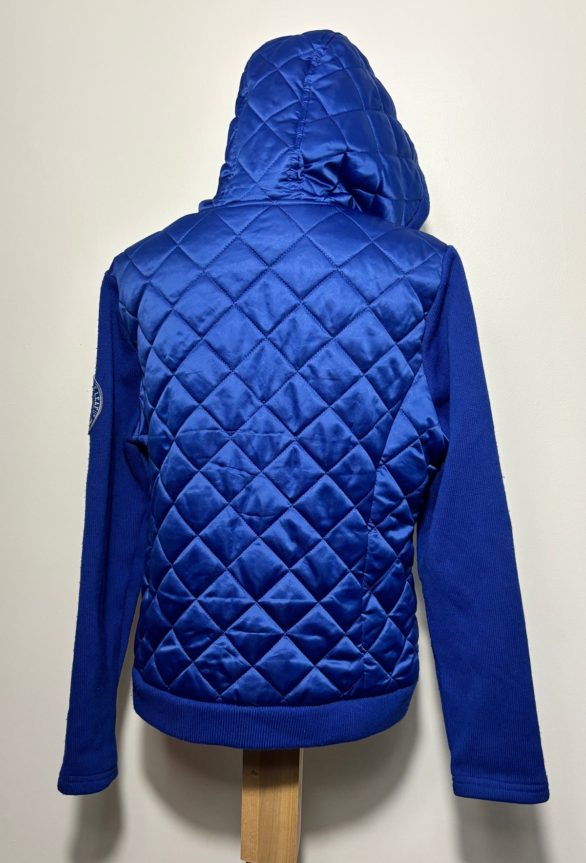 A royal blue quilted hooded jacket displayed on a wooden mannequin stand against a plain off-white wall, showing the back view with diamond quilting and solid blue sleeves.