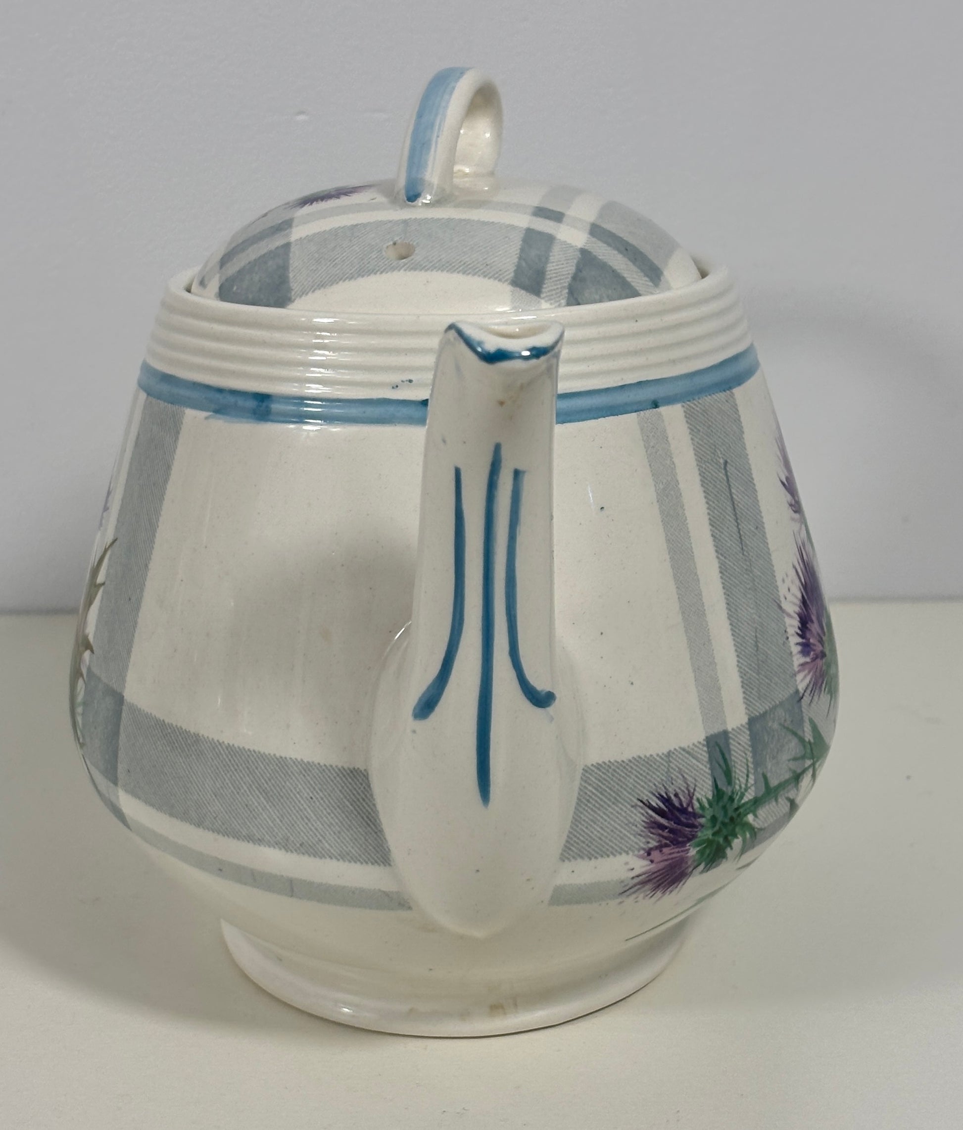 A vintage white ceramic teapot with a lid, decorated with blue stripes, a blue handle accent, and purple thistle floral illustrations on the side.